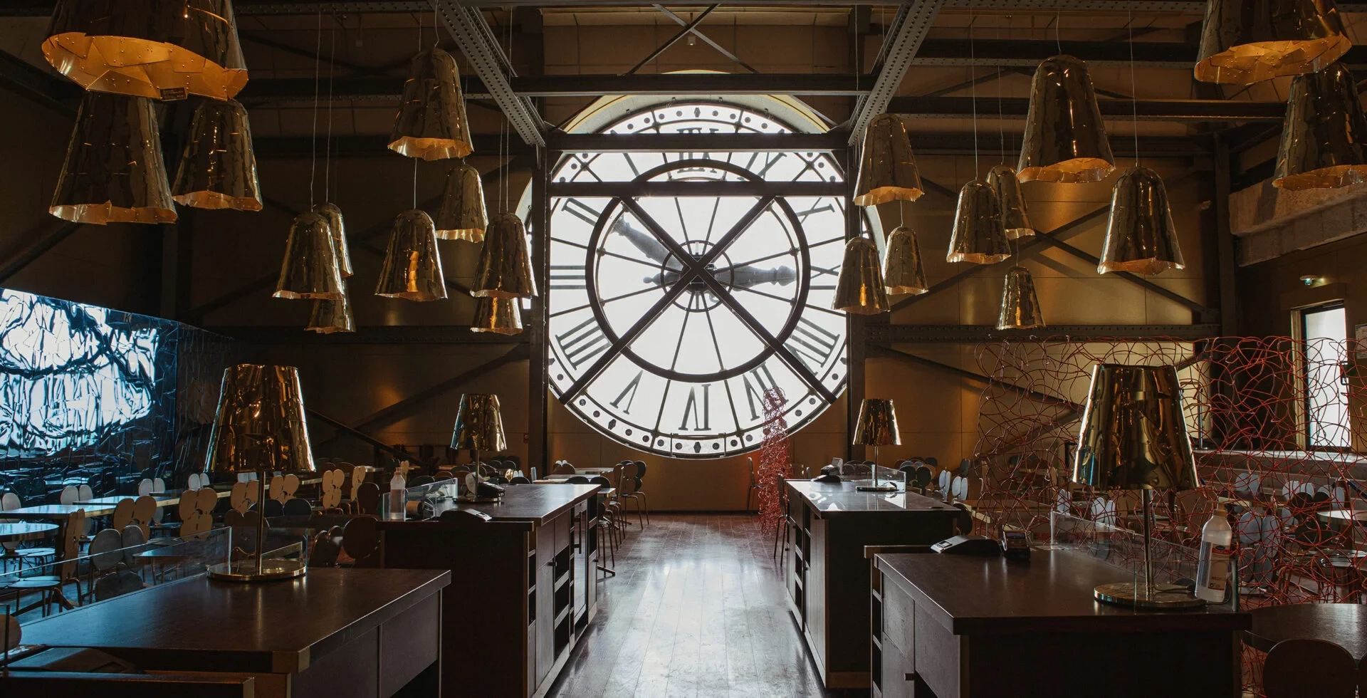 Bir zamanlar Paris'in Orsay Garı'nın bir parçası olan, Belle Époque dönemine ait görkemli iç mekânıyla dikkat çeken Café Campana'dan, Roman rakamlı devasa saat penceresi ve çağdaş altın rengi sarkıt lambalarla aydınlatılan, tarihi dokusu ile modern tasarımı birleştiren, davetkâr bir müze restoranı veya kafe görünümü.