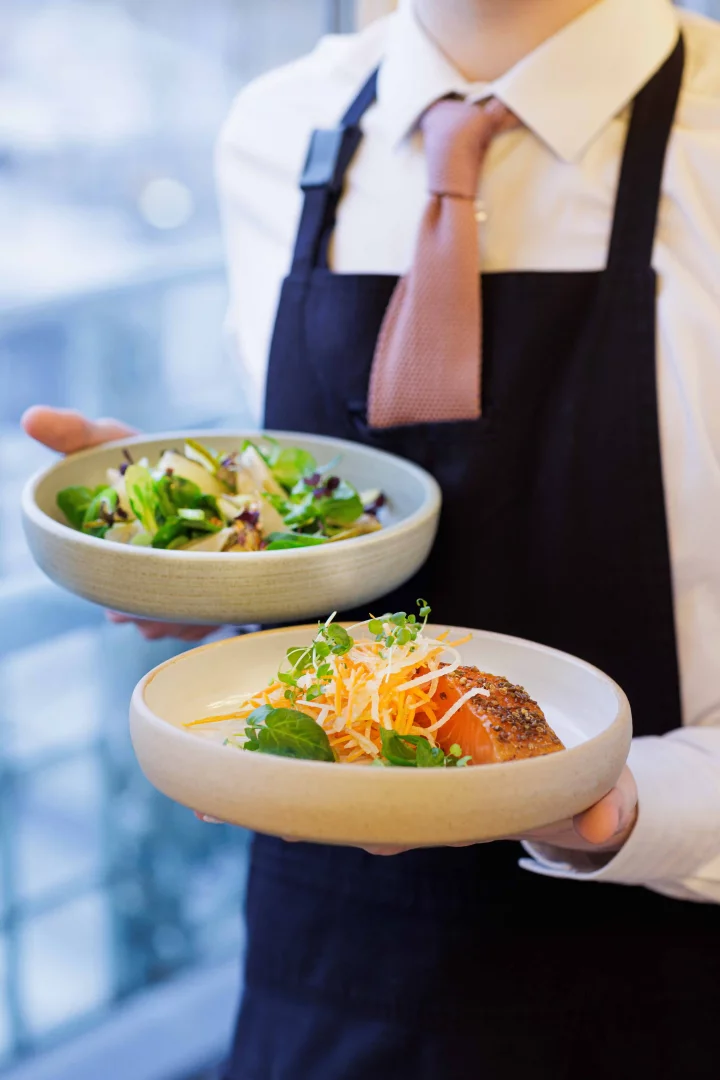 Beyaz gömlekli, kravatlı ve koyu renk önlüklü bir garson, çağdaş müze restoranlarının yükselen gastronomi deneyimini yansıtan, taze yeşilliklerle zenginleştirilmiş bir salata ve özenle hazırlanmış, üzeri rendelenmiş havuç ve mikro yeşilliklerle süslenmiş somon fileto olmak üzere iki sanat eseri niteliğindeki tabağı zarifçe sunuyor.