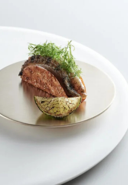 Hong Kong'daki M+ Çağdaş Sanat Müzesi bünyesindeki Michelin yıldızlı Mosu'nun, çıtır kabuklu bir ana yemeği, incecik doğranmış otlar ve desenli bir narenciye dilimiyle sanatsal bir biçimde sunarak, müze restoranlarının artık başlı başına bir seyahat nedeni haline geldiğini gösteren zarif ve davetkar bir görsel.