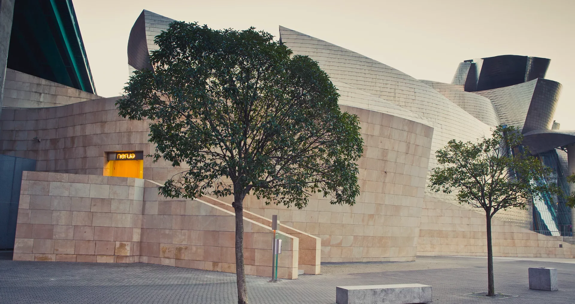 Bilbao'daki Guggenheim Müzesi'nin dalgalı ve fütüristik mimarisi önünde, yemyeşil ağaçların ve Nerua müze restoranının sarı tabelalı girişinin görüldüğü, gastronomi tutkunlarını cezbeden benzersiz bir müze restoranı manzarası.