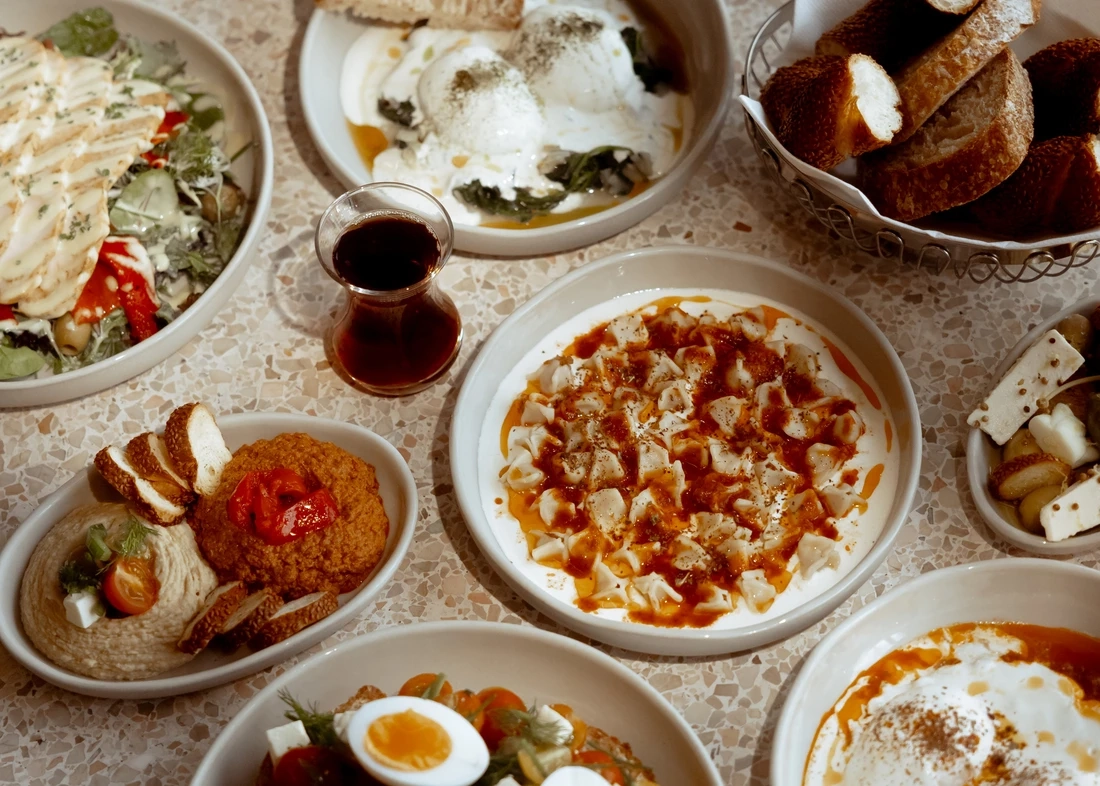 Açık renkli, benekli bir masanın üzerinde, yoğurtlu yumurtalar, mantı, humus, muhammara, çeşitli salatalar, dilimlenmiş simit ve bir bardak demli Türk çayı gibi geleneksel Anadolu lezzetleriyle dolu beyaz kaselerden oluşan zengin bir kahvaltı sofrası, müze restoranlarının başlı başına bir seyahat nedeni olabileceğini vurguluyor.
