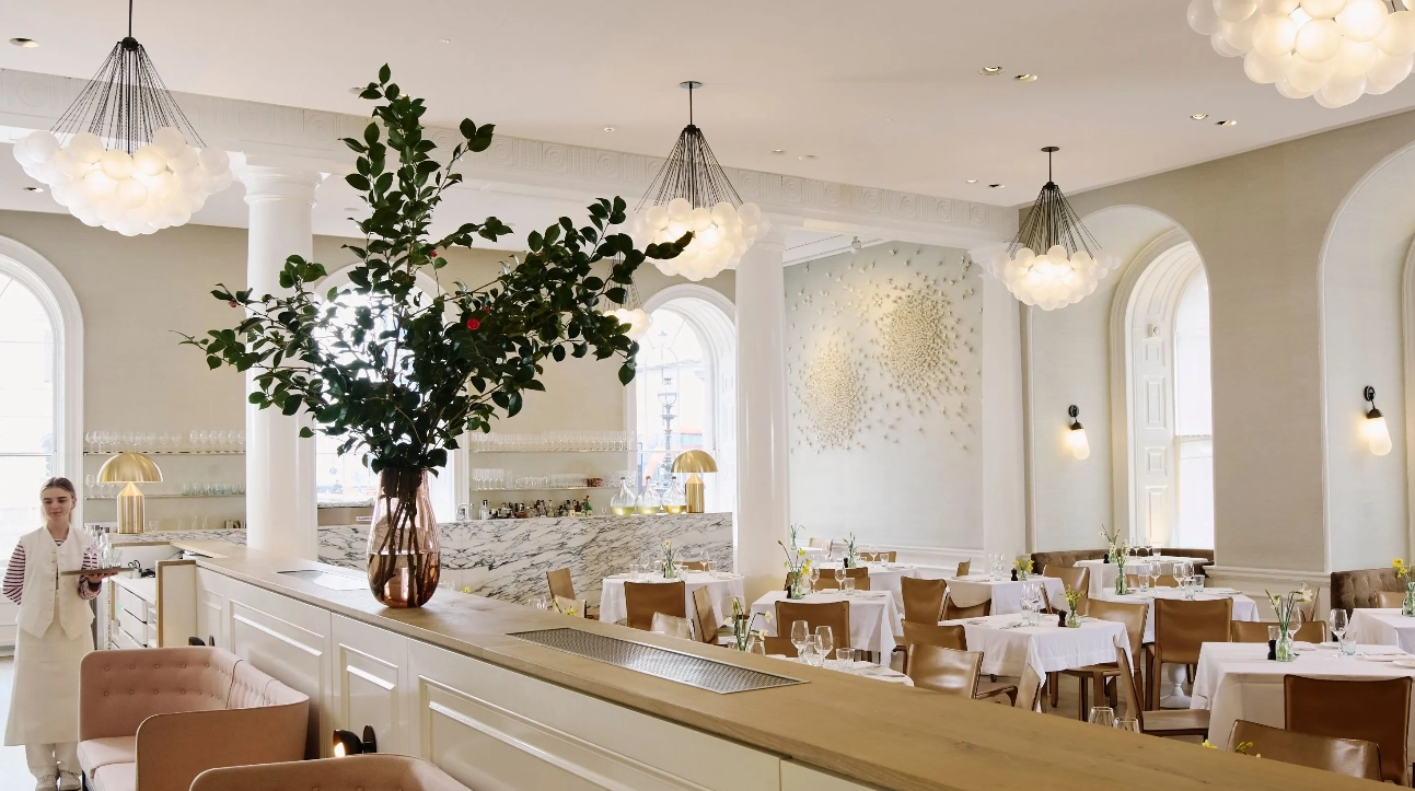 Somerset House'un ferah ve gün ışığıyla dolu, yüksek tavanlı yemek salonunda; tavandan sarkan zarif avizeler, sade dekorasyon ve beyaz örtülü masalar göz alırken, bir görevli müze restoranlarının yükselen gastronomi deneyimini yansıtan bir atmosferde servis yapmaya hazırlanıyor.