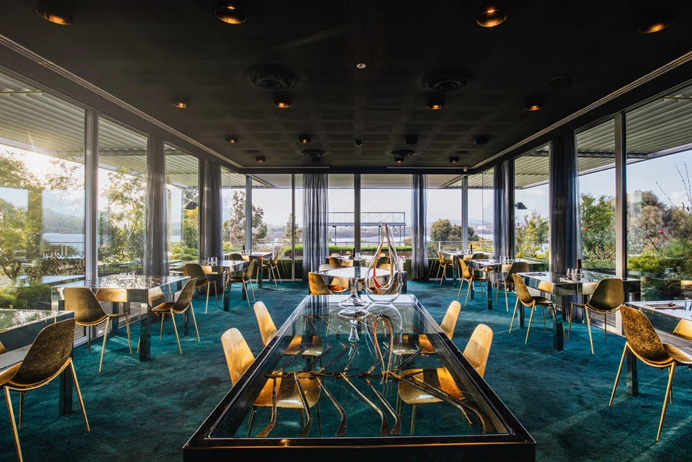 MONA'nın The Source müze restoranında, Derwent Nehri manzaralı geniş cam pencerelerle çevrili şık mekânda, altın rengi sandalyeler ve sanatsal detaylarla süslenmiş cam masalar modern bir gastronomi deneyimi sunuyor.