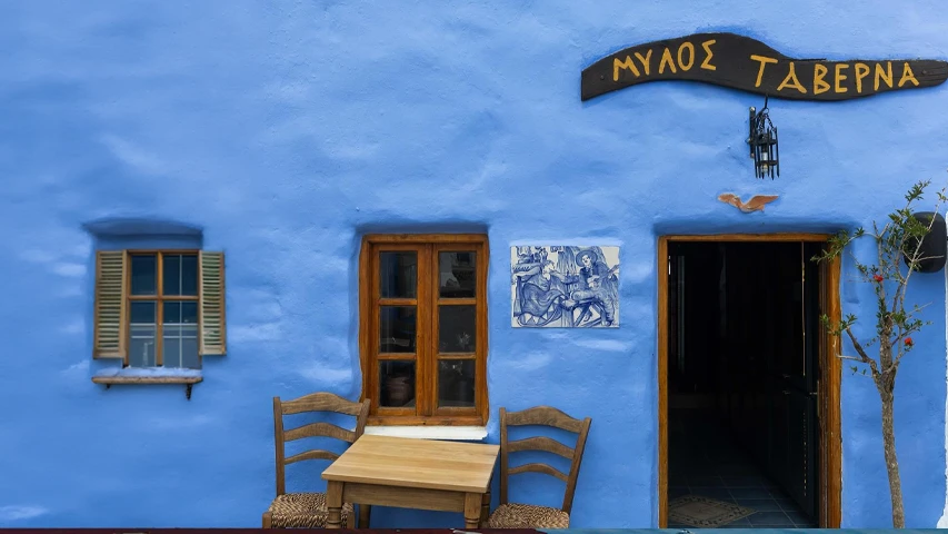 Leros'taki Mylos Taverna tabelasının asılı olduğu canlı mavi bir binanın önünde ahşap pencereler, kapının yanında küçük bir ağaç, dış mekan masa ve sandalyeler ve müzisyenleri gösteren bir seramik pano bulunuyor.