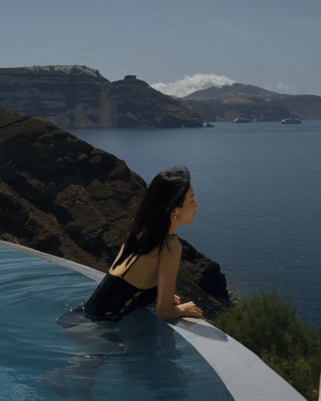 Mystique Otel Santorini'deki lüks sonsuzluk havuzunda, siyah mayolu genç bir kadın Ege Denizi'nin nefes kesici manzarasına, uzaktaki volkanik dağlara ve ufuktaki gemilere doğru dalgınca bakıyor.