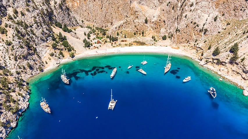 Simi Adası'ndaki Nanou Plajı'nın kuşbakışı görüntüsünde, kayalık yamaçlar arasında uzanan berrak turkuaz sulara sahip gizli bir koyda demirlemiş çok sayıda tekne ve güneşin parladığı sakin bir plaj manzarası yer alıyor.