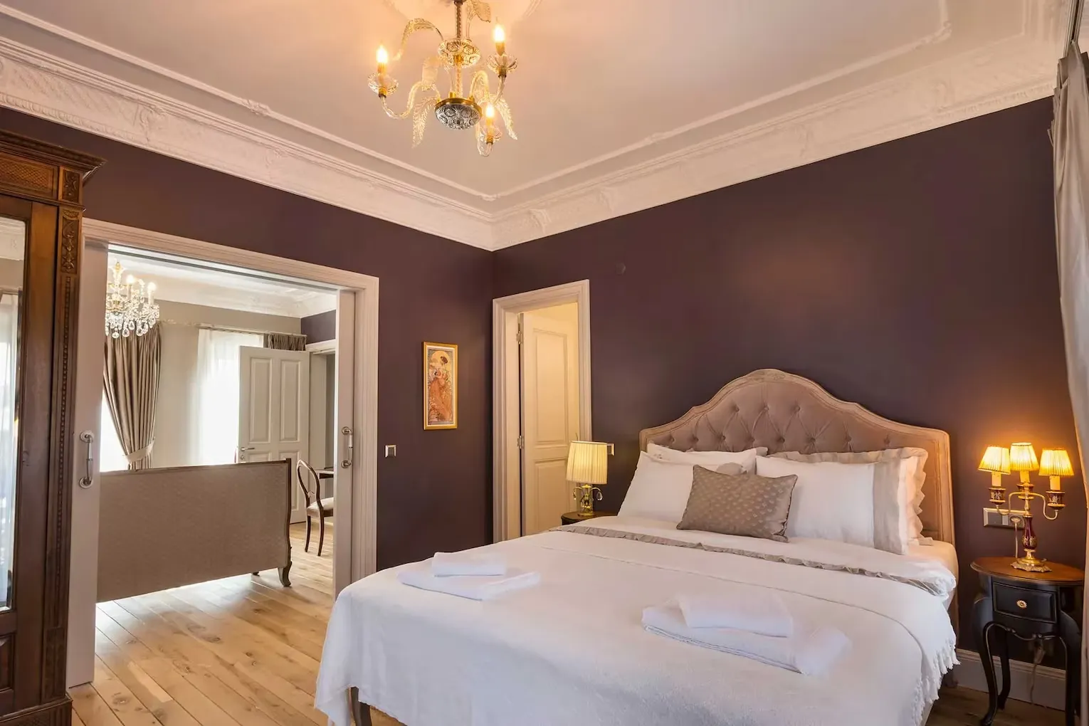 Koyu mor duvarları, beyaz süslemeli tavanı ve altın varaklı avizesiyle dikkat çeken bu şık otel odasında, bej renkli, kabarık başlıklı geniş yatak, beyaz nevresimler ve iki katlanmış havluyla davetkar bir görünüme sahipken, sağ tarafta sıcak ışık yayan üç lambalı bir komodin ve sol tarafta ahşap zeminli, daha aydınlık bir odaya açılan kapı görülüyor.