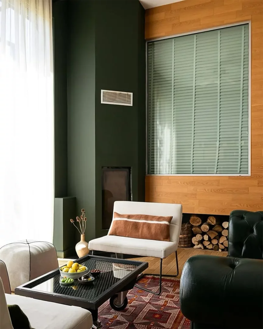 Koyu yeşil duvarlara sahip, modern bir şöminesi ve ahşap panelli penceresi bulunan bu yaşam alanında, krem renkli bir berjer, koyu yeşil deri koltuk, geometrik desenli bir kilim ve üzerinde limonlar ile zeytinlerin olduğu tekerlekli metal sehpa ile sıcak ve şık bir atmosfer oluşturulmuş.