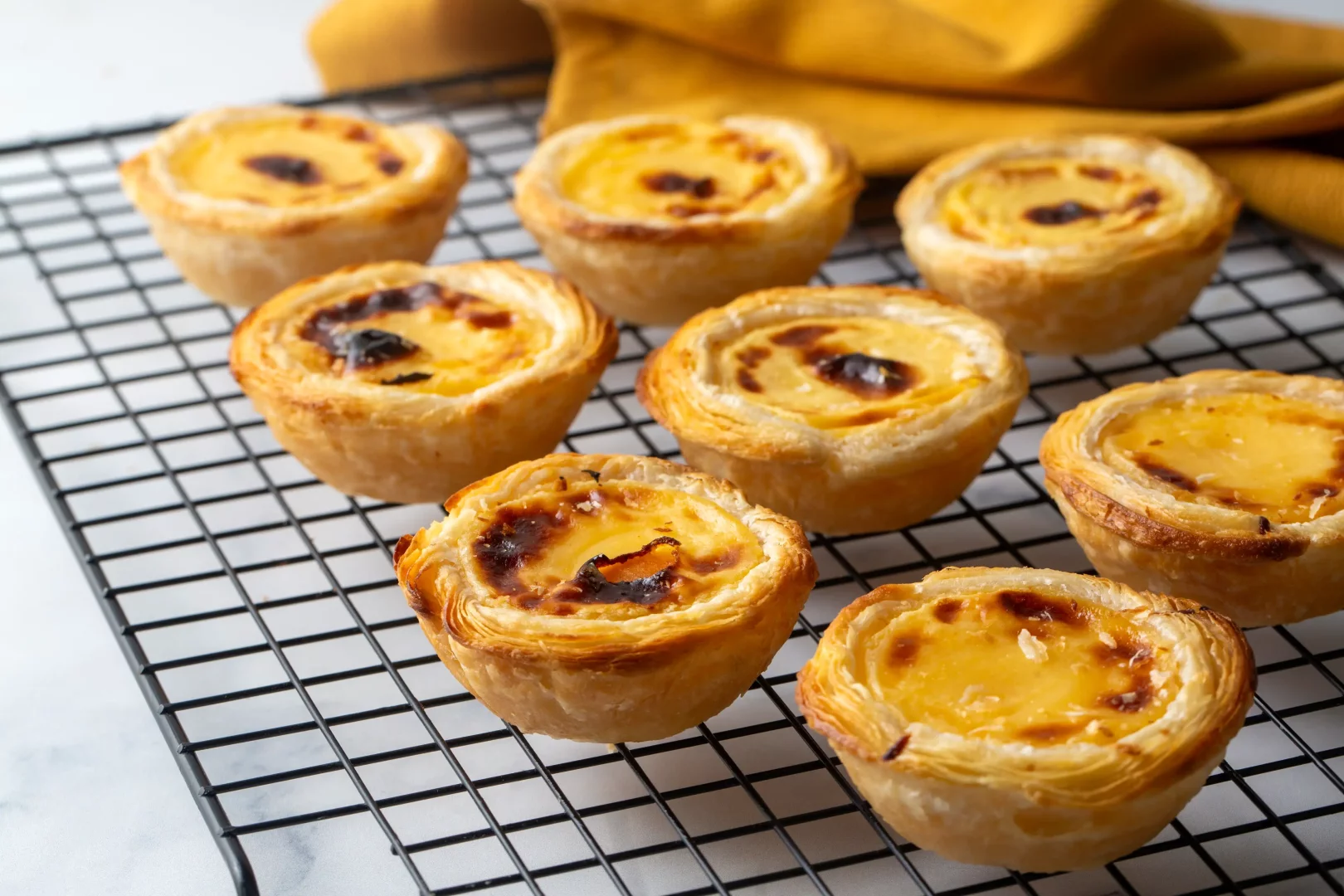 Siyah tel ızgara üzerinde dizilmiş sekiz adet çıtır, katmanlı hamurlu ve üzeri hafifçe karamelize olmuş kremalı sarı muhallebili Portekiz muhallebi tartı (Pastel de Nata) iştah açıcı bir şekilde görülüyor.