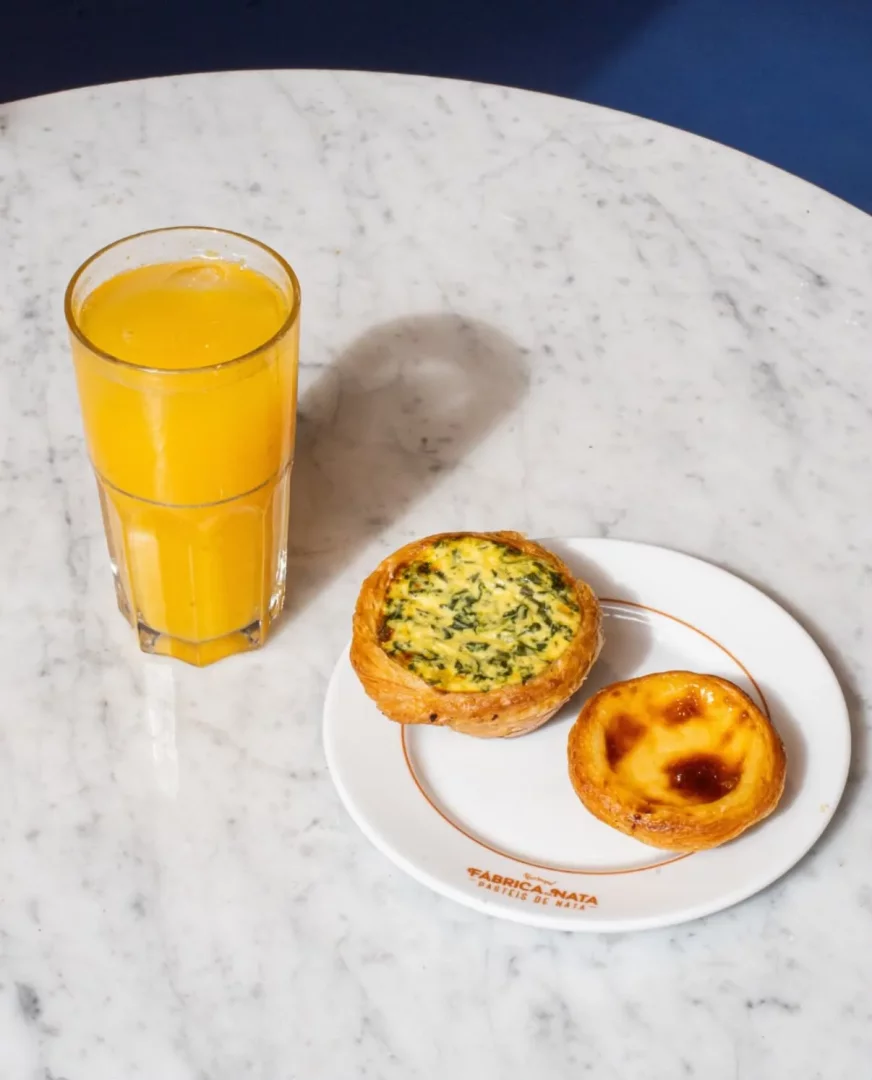 Bir mermer masanın üzerinde, Fábrica de Nata logolu beyaz bir tabakta yanında taze sıkılmış portakal suyu ile servis edilen, Portekiz'in meşhur muhallebili tatlısı pastel de nata ve ıspanaklı tuzlu bir tart.