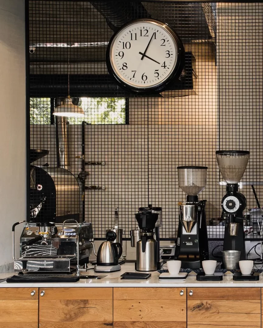 Siyah metal ızgara duvarın ve asılı büyük yuvarlak duvar saatinin önünde, pırıl pırıl bir espresso makinesi, profesyonel iki kahve öğütücü, su ısıtıcısı ve üç küçük beyaz fincanın düzenli bir şekilde yer aldığı mermer bir tezgahta modern bir kahve hazırlık istasyonu ve arka planda kahve kavurma ekipmanları görünüyor.