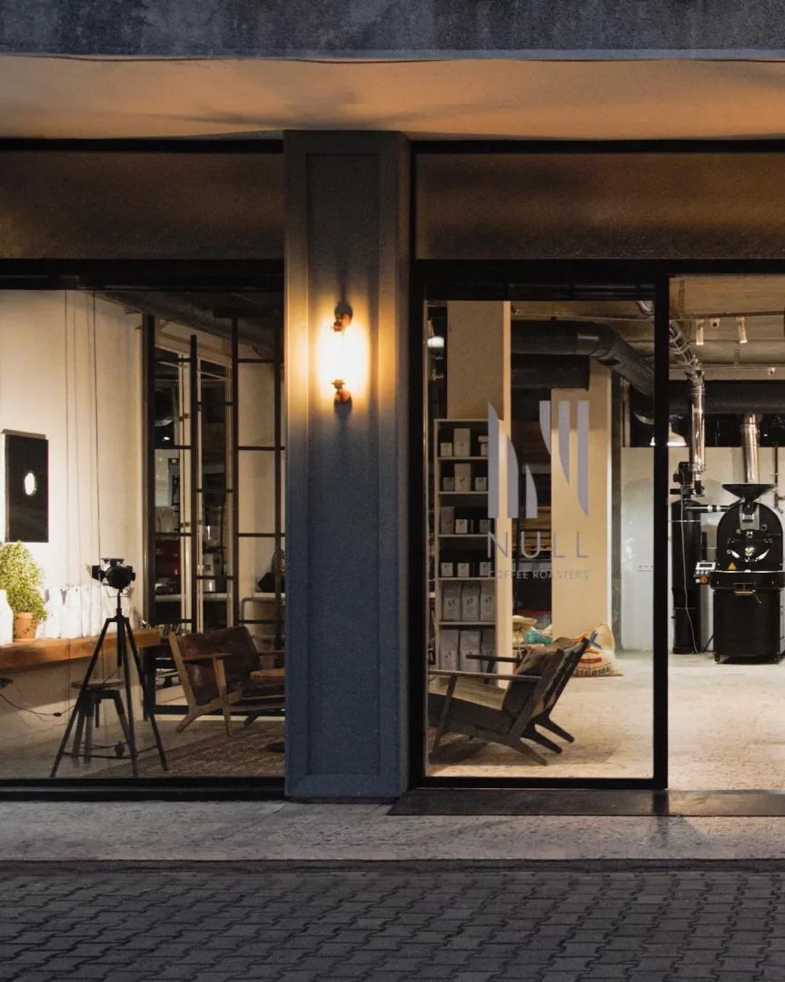 Loş bir akşamda çekilen bu dış mekan fotoğrafında, Null Coffee Roasters'ın sıcak sarı ışıklarla parlayan modern vitrini, içerideki ahşap oturma alanları, kahve rafları ve sağdaki endüstriyel kahve kavurma makinesiyle davetkar bir atmosfer sunuyor.