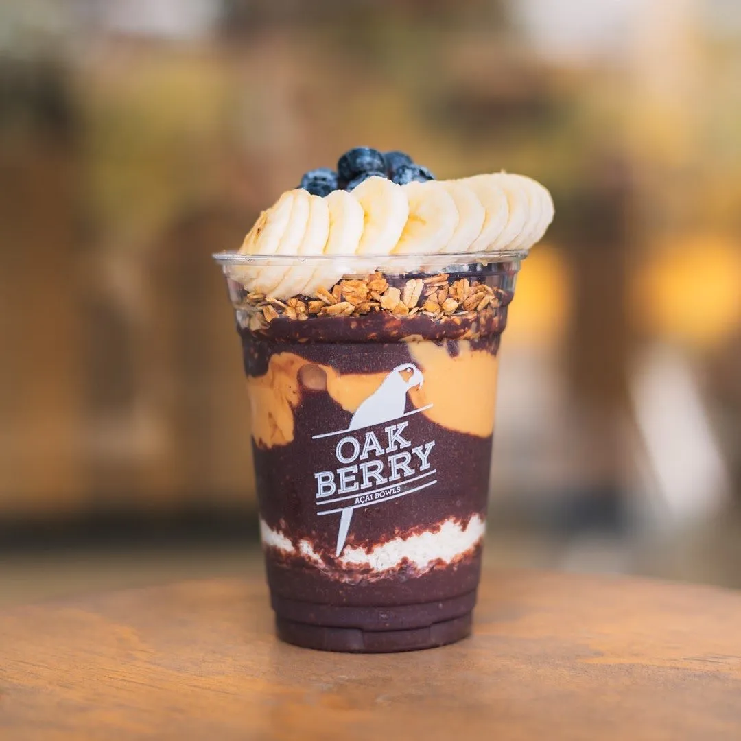 Şeffaf bir Oak Berry Açaí Bowls kupasında, koyu mor açaí, fıstık ezmesi benzeri katmanlar ve krema ile hazırlanan, üzerine granola, muz dilimleri ve yaban mersini eklenmiş lezzetli bir açaí kasesi görüyoruz.