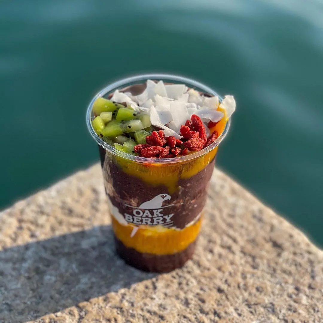 Oakberry logolu şeffaf bir kapta, katmanlı koyu renkli açai, parlak turuncu meyve püresi ile harmanlanmış, üzerinde taze doğranmış kivi, beyaz hindistan cevizi pulları ve kırmızı goji meyvelerinin bulunduğu, pürüzlü bir taş yüzeyin üzerinde duran sağlıklı bir açai kasesi, arka planda hafifçe bulanık deniz veya göl suyu görülüyor.
