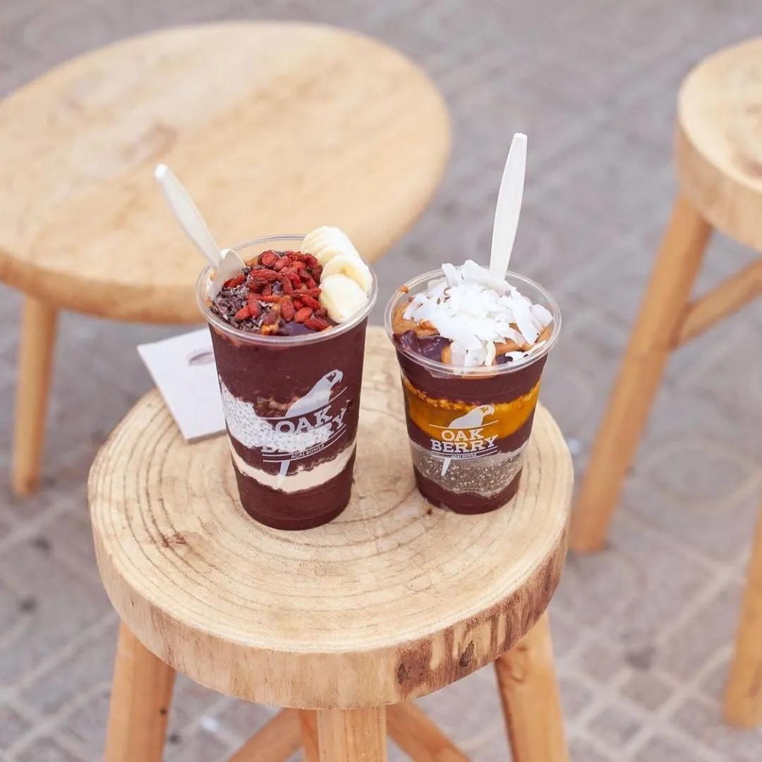 Kesilmiş ağaç kütüğü görünümlü ahşap bir tabure üzerinde duran, Oakberry logosu taşıyan iki şeffaf bardak içinde, katmanlı, rengarenk acai kasesi içecekleri bulunuyor; soldaki goji meyveleri, muz dilimleri ve çikolata parçacıklarıyla, sağdaki ise rendelenmiş hindistan cevizi ve fıstık ezmesiyle süslenmiş.