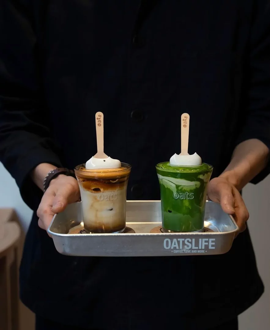 Siyah giyimli birinin elinde, OATSLIFE logosu işlenmiş metal bir tepside, her ikisi de beyaz köpük ve üzerinde #oats yazılı ahşap kaşıklarla servis edilen, biri katmanlı karamel renkli kahve, diğeri ise canlı yeşil renkte matcha olmak üzere iki farklı yulaflı içecek fincanı görünüyor.