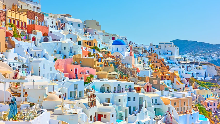 Santorini'nin Oia kasabasının, dik bir yamaçtan masmavi gökyüzü altında sıralanmış, ikonik beyaz ve pastel renkli evleriyle, belirgin mavi kubbeli kiliseler ve teraslarıyla göz kamaştıran panoramik görüntüsü.