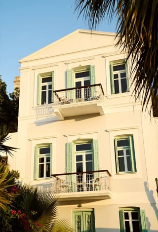 Simi Adası'nda yer alan Old Markets Otel'in nane yeşili panjurlu pencereleri, beyaz korkuluklu balkonları ve ön planda görünen palmiye yapraklarıyla süslenmiş beyaz cephesi, Ege mimarisinin huzurlu atmosferini yansıtıyor.