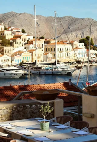 Simi'deki Old Markets Otel'in muhtemelen terasından görünen, iki direkli büyük bir yelkenli teknenin de bulunduğu masmavi limana ve yamaçlardaki renkli geleneksel binalara nazır, üzerinde küçük bir zeytin ağacı olan zarif bir açık hava yemek masası.
