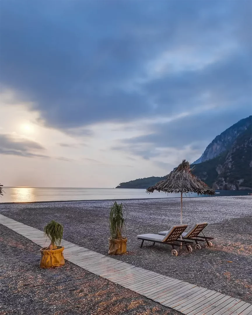 Olympos Lodge'un çakıllı sahilinde, gün batımının yumuşak ışığı altında uzanan ahşap bir yürüyüş yolu, yanında hasır şemsiye ve şezlonglar, durgun deniz ve arkada yükselen dağlarla huzurlu bir manzara.