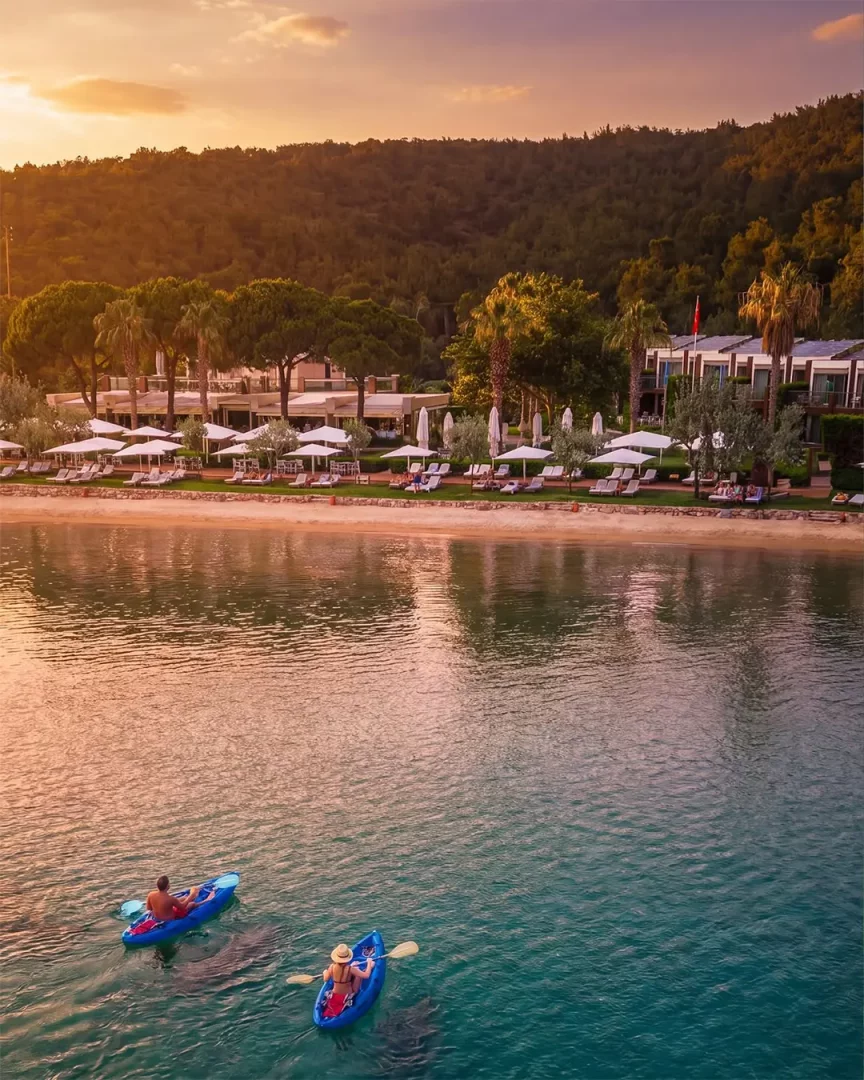 Berrak turkuaz sularda kano yapan iki kişinin görüldüğü bu huzurlu manzarada, Cunda Adası'ndaki Ortunç Otel'in şemsiyeli plajı ve yemyeşil ağaçları gün batımının sıcak renkleriyle büyüleyici bir atmosfer sunuyor.