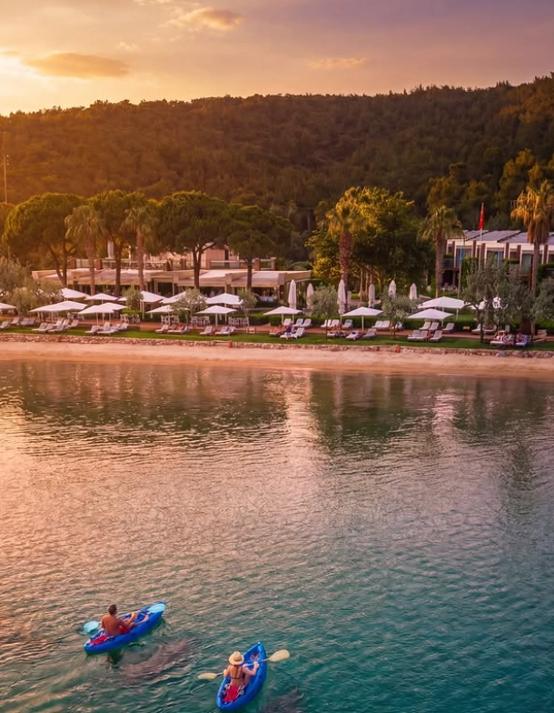 Ortunç Hotel'in berrak turkuaz sularında gün batımı ışıkları altında kayıklarla kürek çeken iki kişinin huzurlu anı, şezlonglar ve beyaz şemsiyelerle dolu sahil şeridi ve yemyeşil ağaçlarla çevrili otel binasıyla Ege'nin mavi yolculuk tutkusunu yansıtıyor.