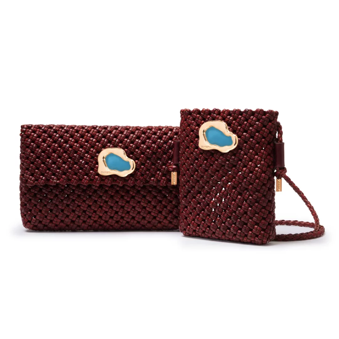 Beyaz bir arka plan üzerinde zarifçe duran, bordo tonlarında, ince işçilikli örgülü deriden yapılmış iki adet Otrera çantası: Soldaki daha büyük olan Iris örgülü clutch, sağdaki ise daha küçük, uyumlu bir çapraz askılı çanta olup her iki çanta da parlak altın rengi, doğal şekilli bir çerçeve içerisine yerleştirilmiş turkuaz bir taş detayıyla süslenmiştir.