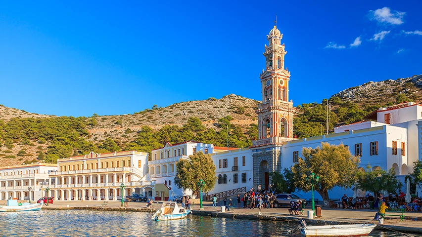 Simi Adası'ndaki Panormitis Manastırı'nın deniz kenarından çekilmiş, görkemli çan kulesi, beyaz binaları, önünde teknelerle dolu masmavi suları ve arka planda yeşil tepeleri gösteren güneşli bir manzara.