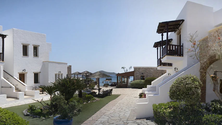 Beyaz badanalı, Ege mimarisindeki binaları, yemyeşil bahçeleri ve taş döşeli yolları ile öne çıkan Panteli Beach Hotel'in güneşli bir dış görünümü, arka planda şemsiyeli plajı ve masmavi deniziyle huzurlu bir atmosfer sunuyor.