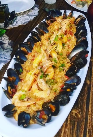 Simi'deki Pantelis Restaurant'ın özel menüsünden, ahşap bir masa üzerinde, bol karidesli ve etrafı siyah kabuklu midyelerle dizili, üzerine taze maydanoz serpilmiş iştah açıcı deniz mahsulleri pilavı, büyük, oval beyaz bir servis tabağında sunuluyor.