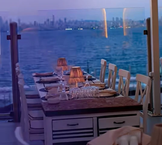 Kınalıada'daki Paros Restoran'ın cam bariyerle çevrili deniz manzaralı terasında, loş ışıklı masa lambalarıyla aydınlatılan, tabaklar ve bardaklarla özenle donatılmış uzun bir masa, karşı kıyıda ışıl ışıl parlayan şehrin panoramik silüeti eşliğinde büyüleyici bir akşam yemeği deneyimi sunuyor.