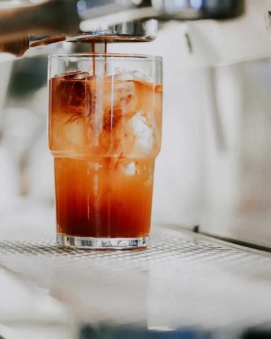 Bir espresso makinesinden taze demlenmiş yoğun espresso, içi buz dolu şeffaf bir bardağa zarifçe süzülerek, alttaki açık renkli buzlu kahve ile mükemmel bir uyum yakalıyor ve özel bir soğuk kahve deneyimi sunuyor.