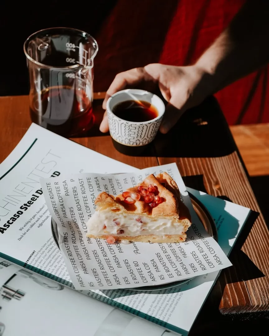 Güneş ışınlarının vurduğu ahşap bir masada, üzerinde Parsa Coffee Roasters logosu bulunan kağıt üzerine konulmuş nar taneli, kremalı bir dilim cheesecake ile birlikte, ölçülü cam bir kapta ve desenli bir fincanda sunulan taze demlenmiş kahveler, keyifli bir molayı çağrıştırıyor.