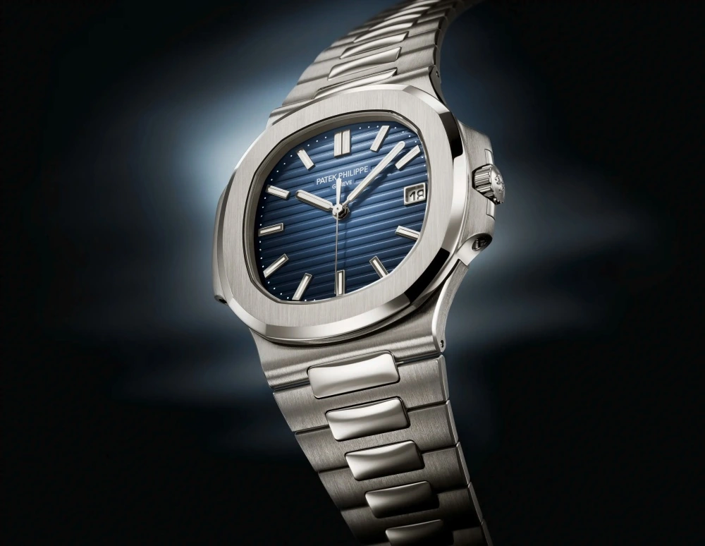 Yakın çekimde, ışıklı koyu bir arka plan üzerinde, beyaz altın kasalı ve bilezikli, ikonik yatay çizgili lacivert kadranıyla zarif Patek Philippe Nautilus 5811 kol saati, 3 yönünde bir tarih penceresiyle dikkat çekiyor.