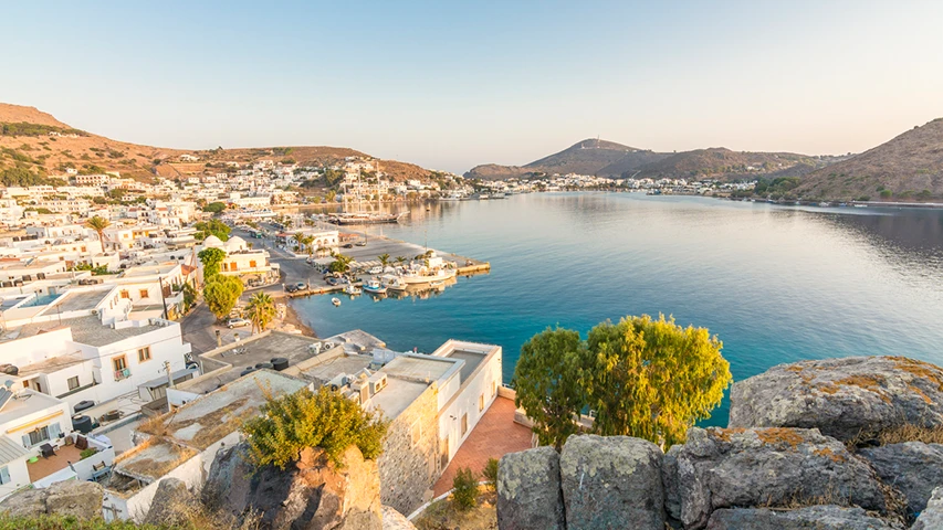 Yemyeşil tepelerle çevrili, beyaza boyalı binalarıyla tipik bir Patmos Adası yerleşimi ve teknelerle dolu sakin bir liman koyunun geniş açılı görünümü, Patmos Adası'na nasıl gidileceğini düşünenler için davetkar bir atmosfer sunuyor.