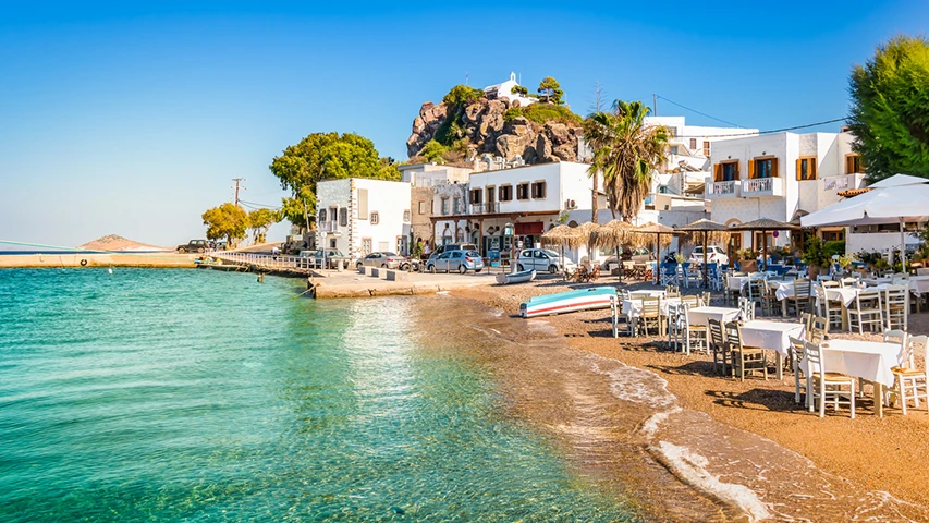 Patmos Adası'nda yaz mevsiminde, berrak turkuaz renkteki denizin kenarında uzanan daracık kumsalda dizili, beyaz masa ve sandalyeleriyle misafirlerini ağırlamaya hazır, şemsiyelerle gölgelenmiş açık hava restoranlarının ve beyaz badanalı binaların yer aldığı, kayalık bir tepenin üzerinde ise küçük bir kilisenin yükseldiği hareketli bir sahil kasabası görünümü.