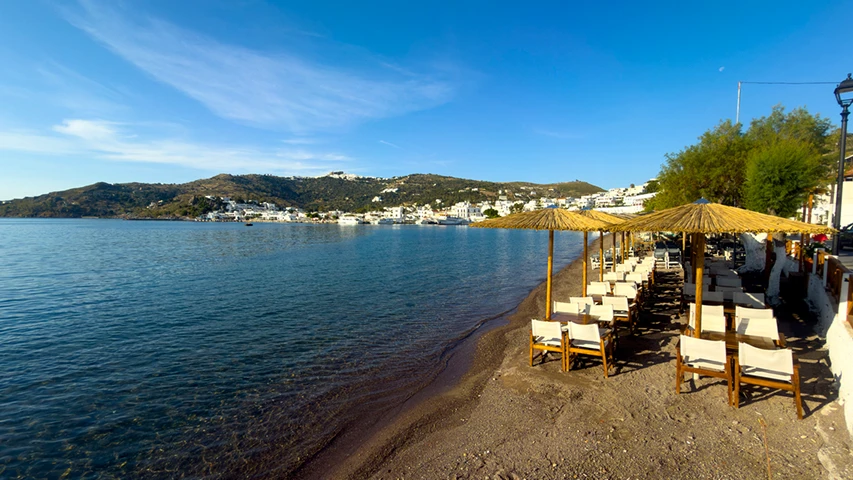 Patmos Adası'ndaki bir plajda, berrak denize sıfır konumlanmış, sıralı hasır şemsiyelerin altında boş masa ve sandalyelerin bulunduğu sakin bir sahil kafesi ile karşı kıyıda beyaz evlerin sıralandığı yemyeşil tepelerin görüldüğü güneşli bir manzara.