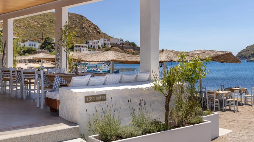 Plessis Taverna tabelalı bu Patmos adası sahil restoranında, bembeyaz ahşap sandalyeler ve kareli masa örtülü masalar, çizgili minderli taş oturma alanı, uzaklarda demirli teknelerin görüldüğü masmavi deniz ve yamaçlardaki beyaz evlerin eşlik ettiği güneşli, huzurlu bir Akdeniz ambiyansı.