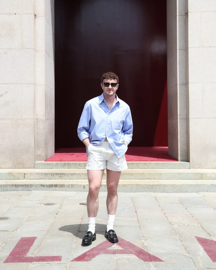 Paul Mescal'ın Milan Moda Haftası kapsamında, güneşli bir günde, üzerinde kırmızı LA harfleri olan taş zeminde durup kameraya baktığı bir an; açık mavi gömleği, çizgili şortu, beyaz çorapları ve metal tokalı siyah Gucci makosen ayakkabılarıyla tamamladığı stil.