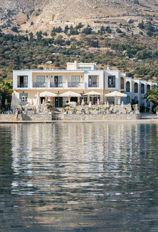 Simi Adası'nda bulunan Pedi Beach Hotel, denizin sakin yansımalı sularına nazır, zemin katında beyaz şemsiyelerle donatılmış açık hava restoranı bulunan modern mimarili bir binadır ve arkasında kayalık, yeşil dağlarla çevrili bir koy manzarası sunar.