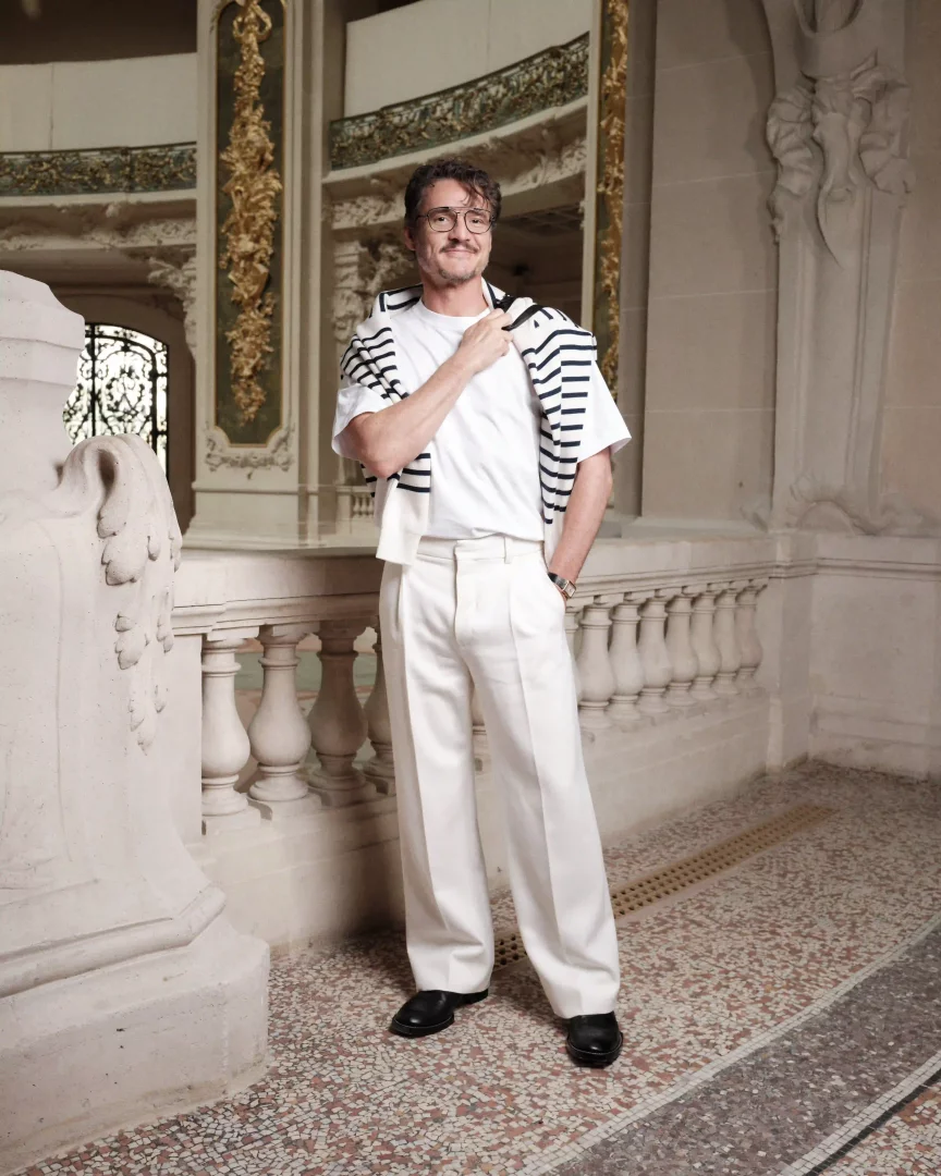 Pedro Pascal, Paris Moda Haftası etkinliğinde beyaz tişört ve bol pantolon üzerine siyah beyaz çizgili bir süveteri omuzlarına atmış, gözlükleriyle süslü bir iç mekanda kameraya hafifçe gülümseyerek poz veriyor.