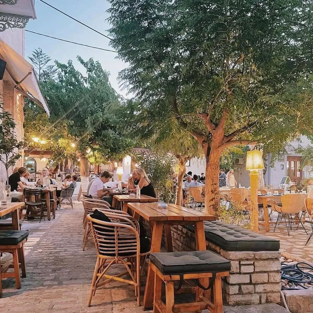 Ağaçların gölgesindeki Pergamonto adlı açık hava restoranında, ahşap masalarda ve hasır sandalyelerde oturan çok sayıda misafirin, asılı ışıklandırmaların yarattığı sıcak bir atmosferde sohbet edip keyifli zaman geçirdiği hareketli ve davetkar bir akşam sahnesi.