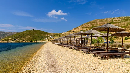 Patmos'taki Petra Plajı'nda, berrak masmavi suyla buluşan çakıllı bir sahilde, arka planda yeşil tepeler ve bulutsuz mavi gökyüzü altında uzanan, ahşap şezlonglarla dolu sıra sıra hasır plaj şemsiyeleri görülüyor.