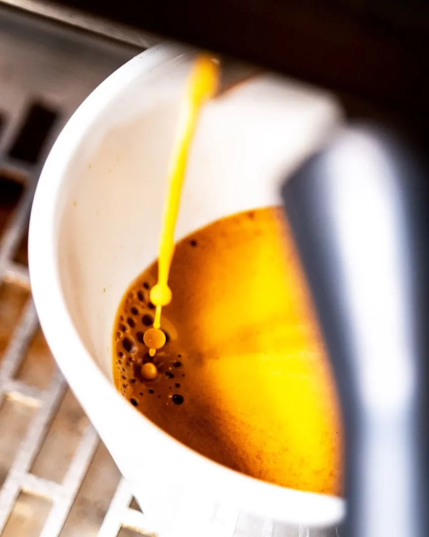 Beyaz bir fincana dökülen zengin amber rengi espresso, koyu kahverengi kahvenin üzerinde altın rengi köpükler (krema) oluştururken, sağda bir kahve makinesinin metalik kısmı ve arka planda bulanık bir desen görülüyor.