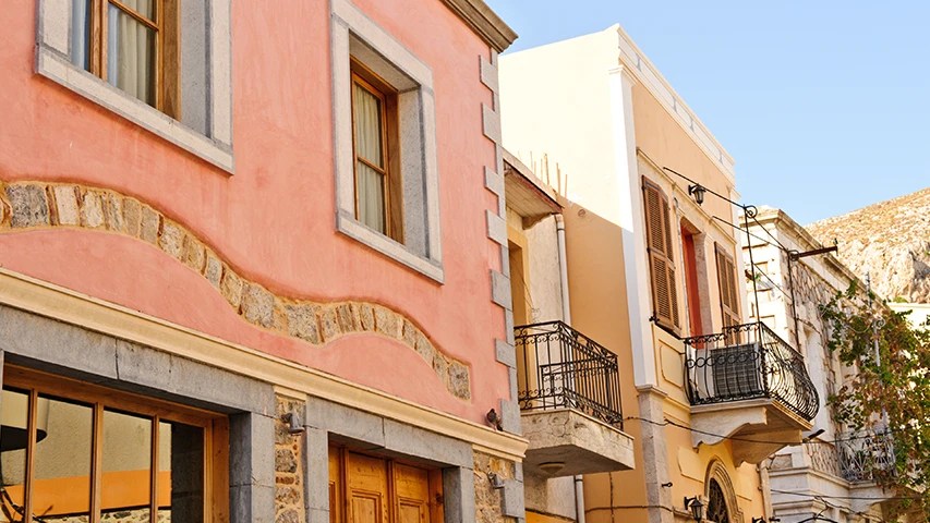 Kalimnos'taki Pothia sokaklarından bir görünüm sunan bu karede, pembe boyalı bir binanın dalgalı taş süslemeleri ve pencereleri, bitişiğindeki ferforje balkonlu ve ahşap panjurlu bej renkli evlerle Akdeniz mimarisinin canlı örneklerini oluşturuyor.