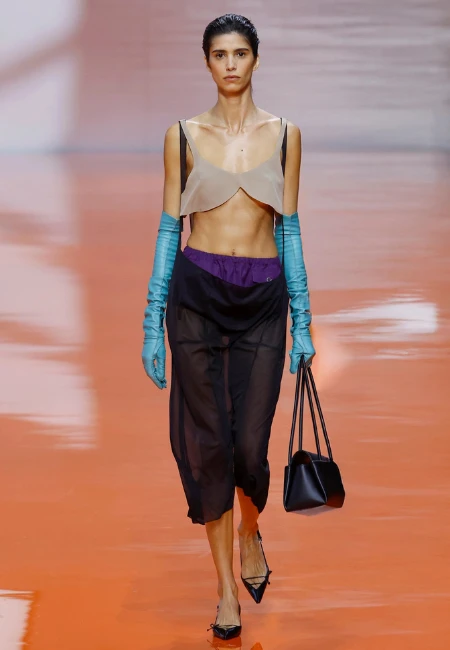 Prada 2026 İlkbahar-Yaz podyumunda, bej bralet, turkuaz uzun eldivenler, mor iç şortun üzerindeki siyah transparan düşük bel midi etek, elde siyah çanta ve topuklu ayakkabılarla yürüyen bir model, yaz etek trendlerini yansıtan deneysel katmanlı bir görünüm sergiliyor.