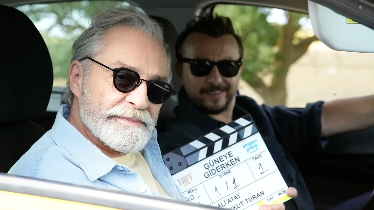 Yeni Amazon Prime filmi Güneye Giderken çekimleri esnasında, Haluk Bilginer güneş gözlüklü ve beyaz sakallarıyla aracın ön koltuğundan doğrudan kameraya bakarken, arka planda yönetmen Ali Atay gülümseyerek üzerinde filmin adının yazılı olduğu bir klaket tutuyor.