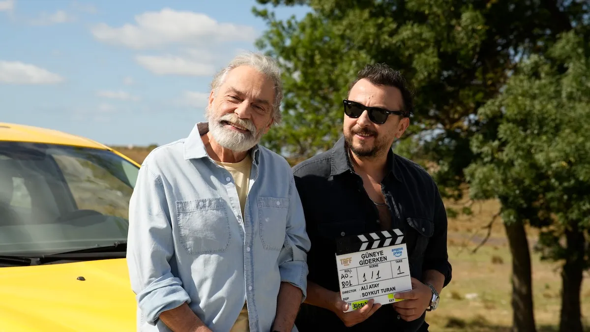 Güneye Giderken filminin çekimleri sırasında, sakallı, yaşlı bir sanatçı ve güneş gözlüklü genç bir ekip üyesi, elinde Güneye Giderken yazan bir klaketle neşeyle poz veriyor, arka planda parlak sarı bir araba ve yeşil ağaçlar açık hava seti atmosferini tamamlıyor.