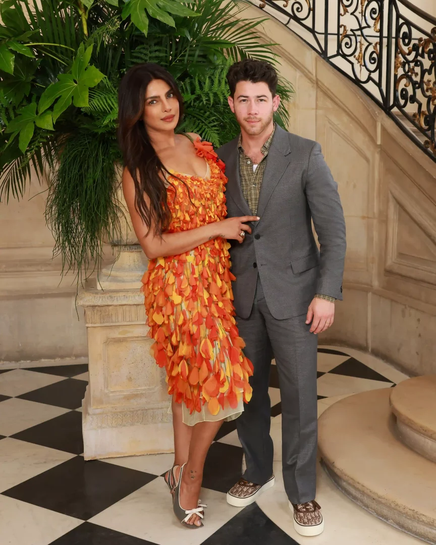Priyanka Chopra Jonas ve Nick Jonas, Paris Haute Couture Moda Haftası kapsamında Dior defilesine katılarak fuaye alanında poz veriyor; Priyanka turuncu tonlarda yaprak aplikeli şık bir elbise giyerken, Nick ise gri bir takım elbise ve desenli ayakkabılarıyla modern couture stilini yansıtıyor.