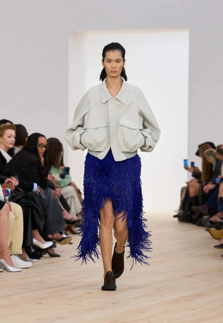 Loewe'nin İlkbahar-Yaz 2026 defilesinde, açık gri oversize bir ceketle tamamlanmış, canlı elektrik mavisi tonlarında, diz hizasından başlayıp arkaya doğru uzayan, yürüdükçe dinamik bir hareket kazanan yoğun püskül detaylarına sahip asimetrik midi bir etek giyen model, 2026 yazının dikkat çekici etek trendlerinden fringe detayları ustalıkla sunuyor.